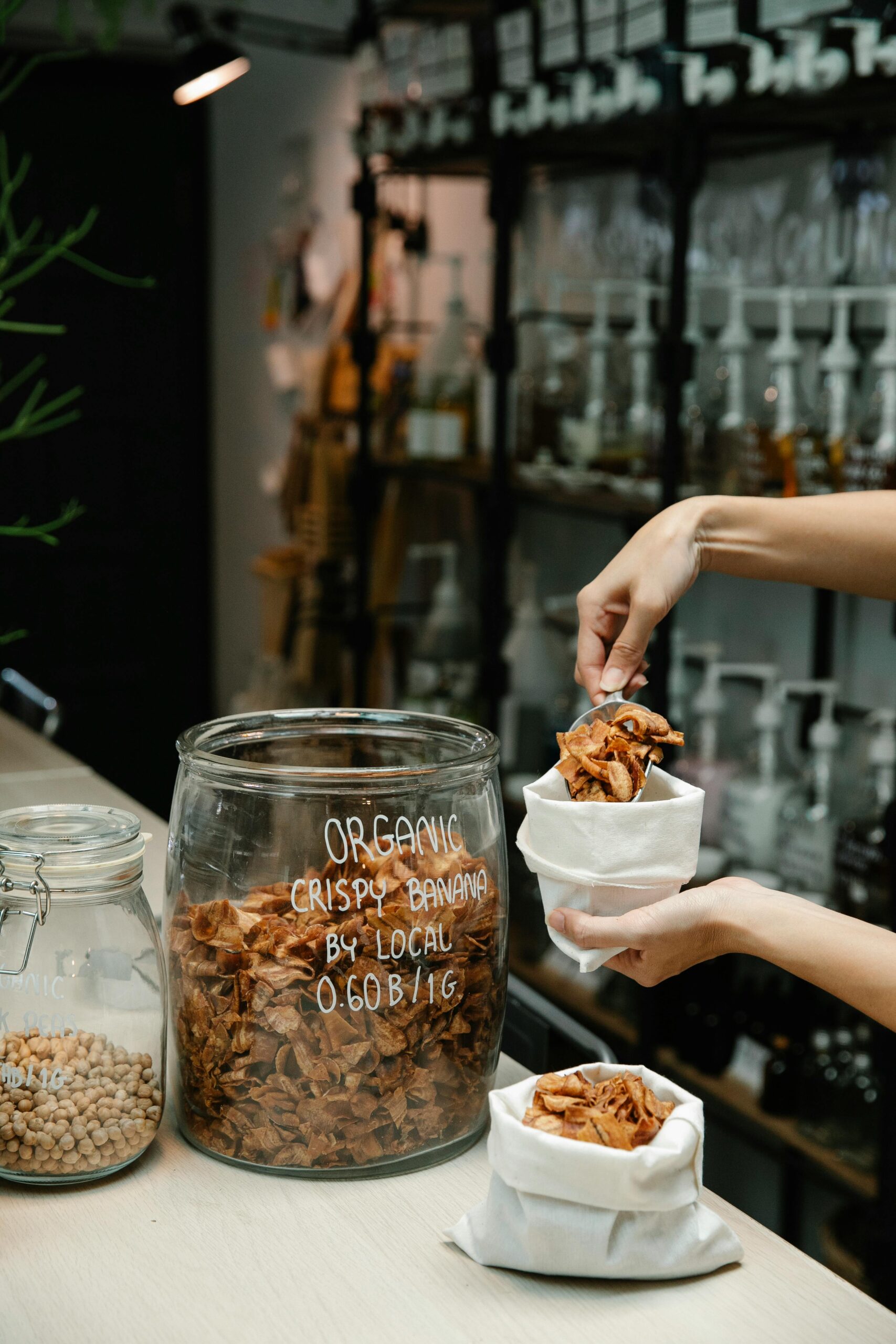 Hand scooping organic crispy banana chips into a paper bag at a local shop.